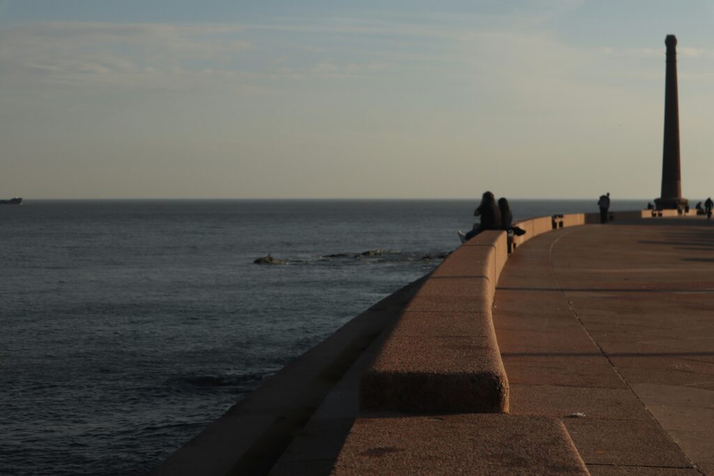 Duas pessoas estão sentadas em uma saliência de pedra perto do mar em Montevidéu, com figuras distantes caminhando pelo caminho curvo sob uma estrutura alta e estreita.