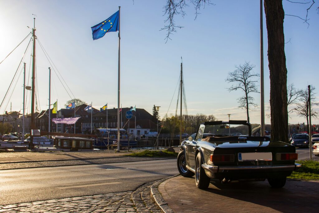 Um carro conversível clássico estacionado em Neustadt in Holstein em uma marina sob um céu limpo, com uma bandeira europeia tremulando em um mastro próximo. Representa aluguel de carros na Alemanha.