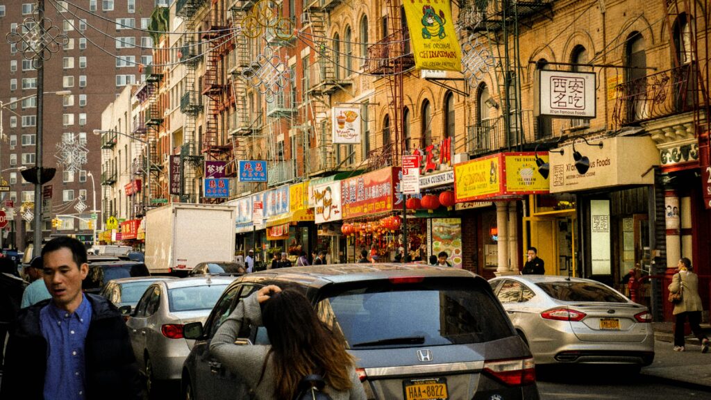 Rua urbana movimentada de Chinatown em New York, com fachadas coloridas, pessoas andando e carros no trânsito. Placas brilhantes são exibidas acima das lojas. Representa aluguel de carros nos Estados Unidos.
