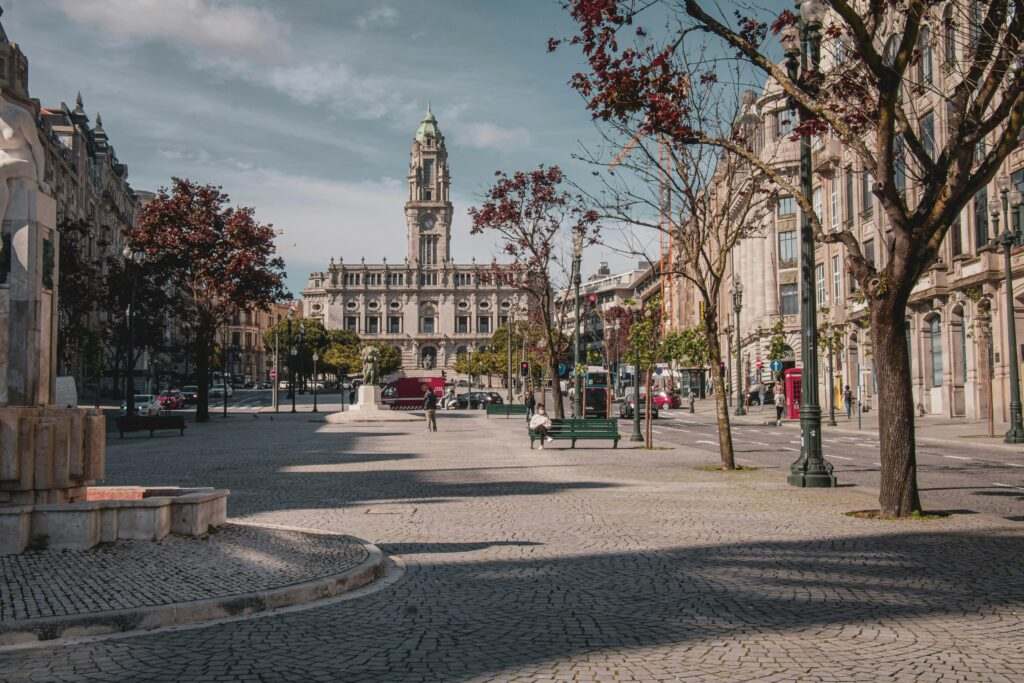 Uma praça ampla e vazia no Porto, com calçada de paralelepípedos, cercada por árvores e prédios, levando a um grande edifício histórico ao fundo.