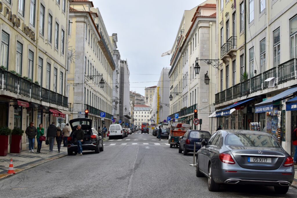 Uma cena de rua de Lisboa com carros estacionados, pessoas caminhando na calçada e prédios de vários andares em ambos os lados. Representa aluguel de carros em Lisboa.