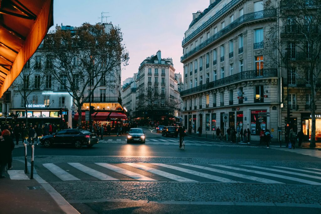 Uma movimentada rua de Paris ao anoitecer, com carros, pedestres e vitrines iluminadas cercadas por arquitetura europeia clássica. Representa aluguel de carros na França.