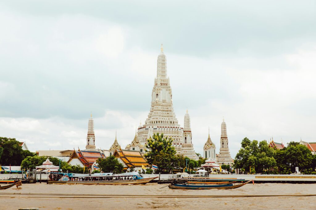 Uma vista do templo Wat Arun em Bangkok, com sua torre central erguendo-se acima de estruturas menores ao redor, vista do outro lado do rio. Representa chip internacional para Bangkok.