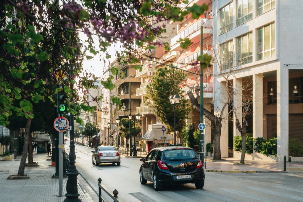 Vista de uma rua em Atenas com carros dirigindo, cercada por prédios e árvores. O semáforo está verde, com um pedestre na calçada esquerda. Representa aluguel de carros em Atenas.