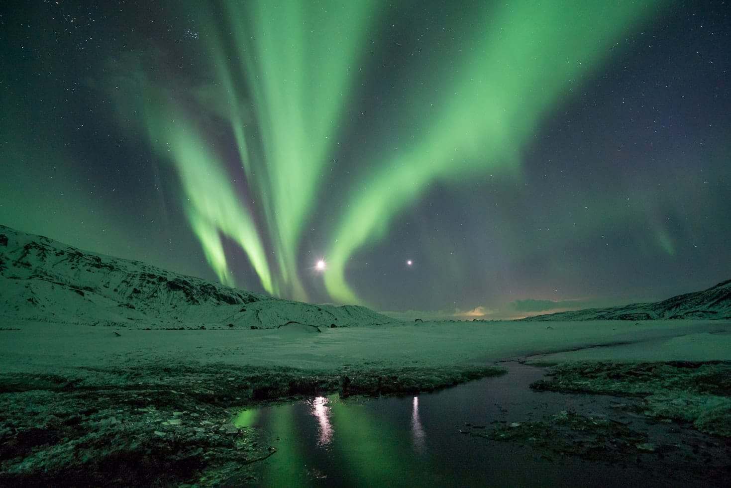 A aurora boreal verde ilumina o céu noturno de Thórsmörk sobre uma paisagem nevada com montanhas e um corpo d'água refletivo. Representa eSIM Islândia