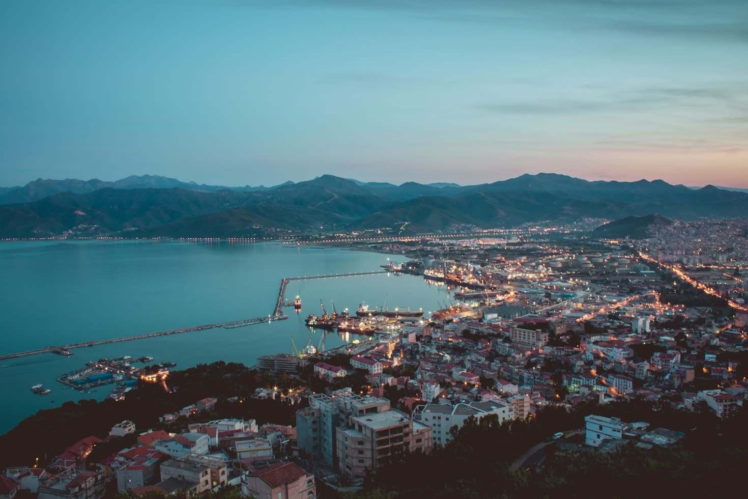Paisagem urbana costeira de Béjaïa ao anoitecer com edifícios iluminados, um porto com barcos e montanhas ao redor sob um céu pouco iluminado. Representa eSIM Argélia.