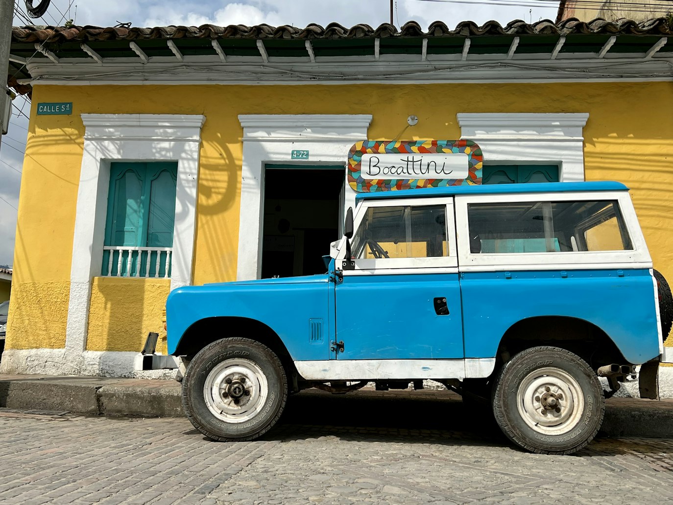 Um SUV azul antigo está estacionado em frente a um prédio amarelo com janelas azul-petróleo em uma rua de paralelepípedos, em Tabio. Representa aluguel de carros na Colômbia.