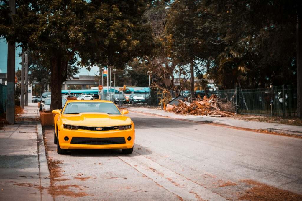 Um Chevrolet Camaro amarelo está estacionado em uma rua ladeada de Miami por árvores e folhas caídas.