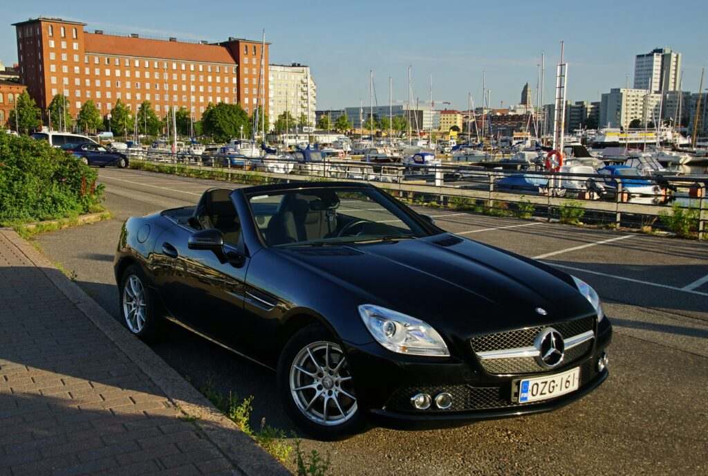 Um Mercedes-Benz conversível preto estacionado perto de uma marina com barcos, tendo como pano de fundo edifícios e um céu limpo, em Helsinki.