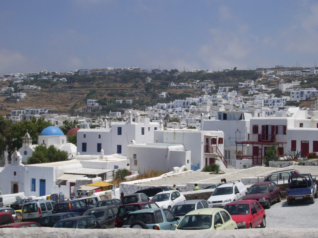 Vista de um estacionamento lotado em Mykonos com carros em primeiro plano e a cidade com prédios brancos e uma estrutura com cúpula azul ao fundo. Representa aluguel de carros em Mykonos.
