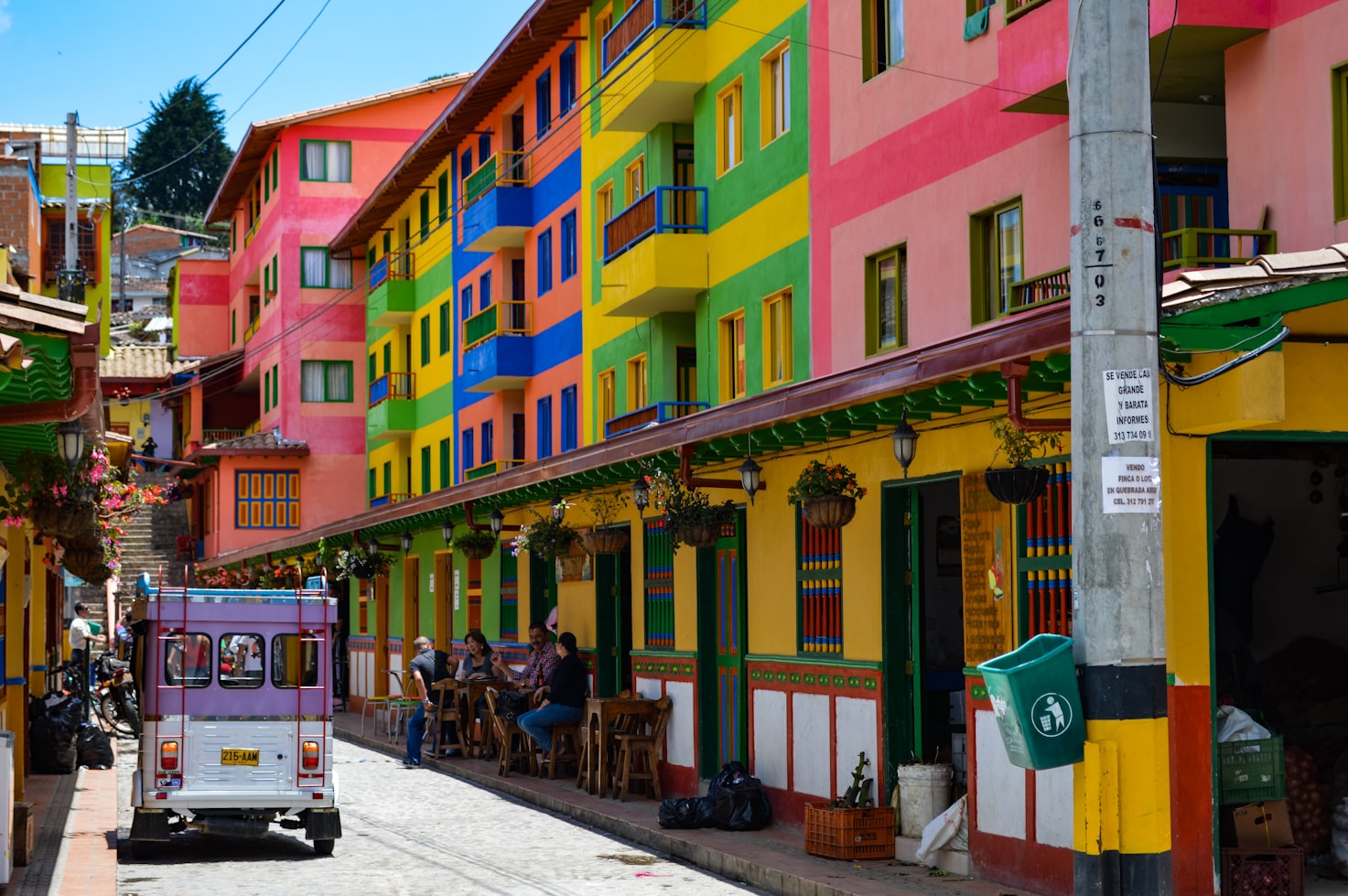 Prédios coloridos alinham uma rua estreita, com um pequeno veículo passando por restaurantes ao ar livre. Plantas penduradas decoram a fachada, na Colômbia. Representa aluguel de carros na Colômbia.