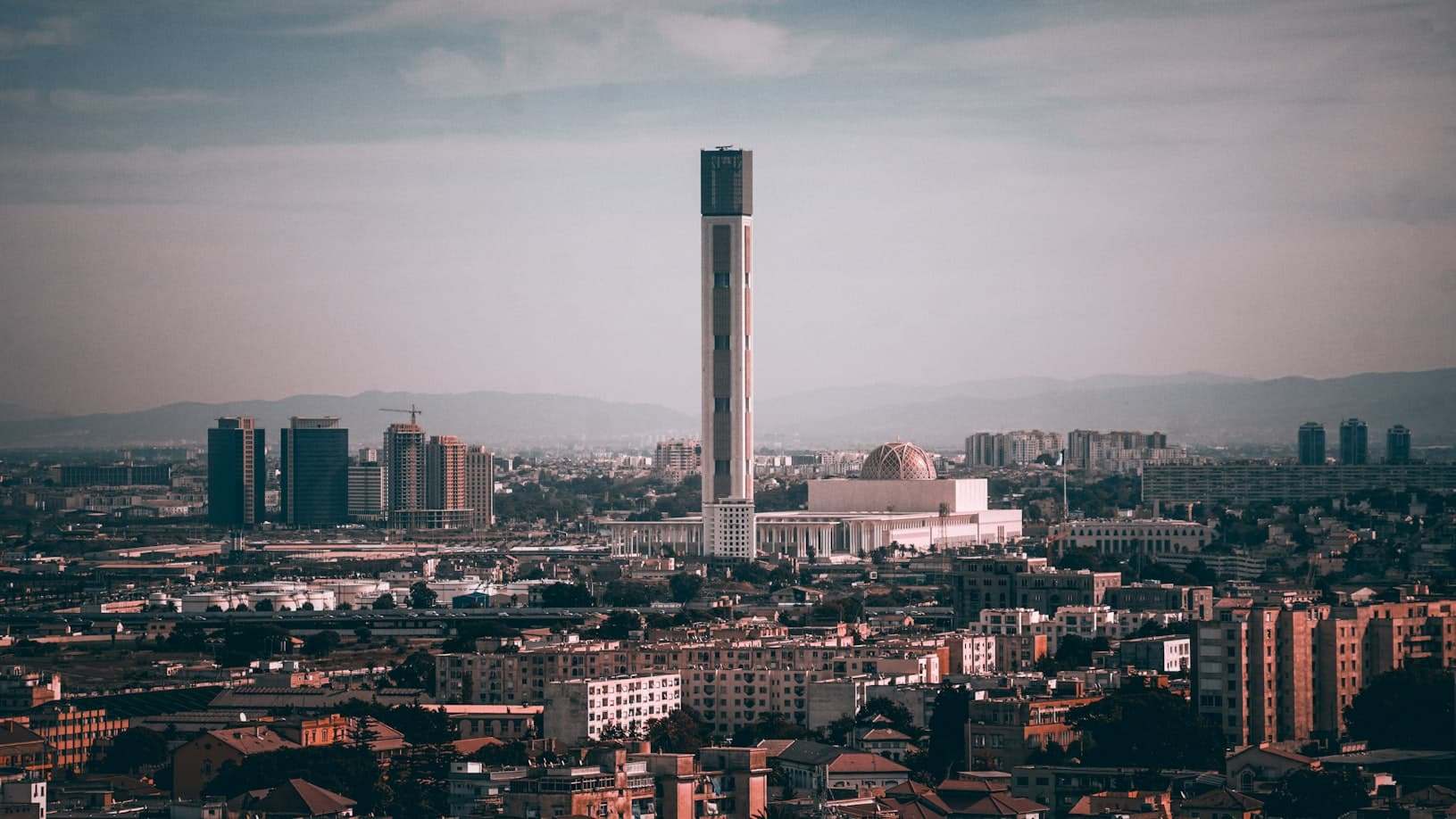 Paisagem urbana da Argélia  com uma torre alta e esbelta e vários prédios ao redor sob um céu nublado. Montanhas distantes são visíveis. Representa eSIM Argélia.