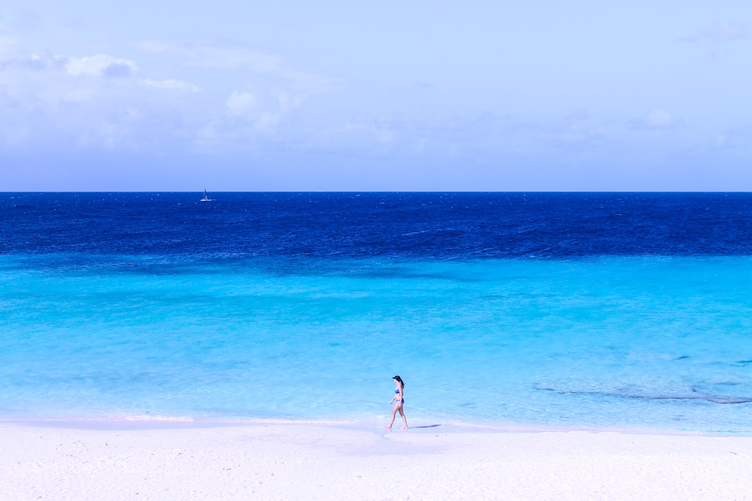 Uma pessoa caminha ao longo de uma praia de areia branca com águas azul-turquesa cristalinas e um veleiro visível à distância sob um céu azul, em Little Curaçao. Representa eSIM Curaçao.