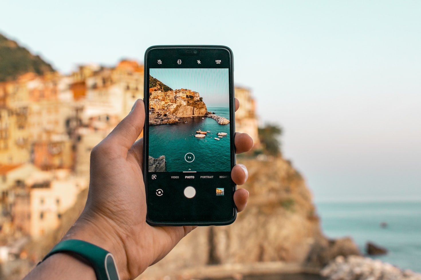 Uma mão segurando um smartphone captura uma cidade costeira com edifícios coloridos e barcos na água.