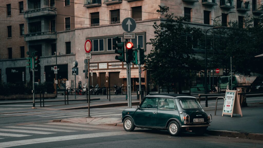Carro verde antigo estacionado em uma esquina com um semáforo vermelho, perto de prédios urbanos e placas de lojas ao fundo, em Milão. Representa aluguel de carros em Milão.