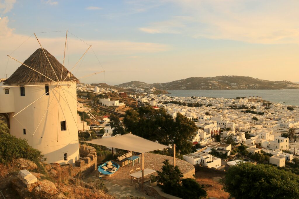 Um moinho de vento tradicional tem vista para Mykonos com edifícios brancos e colinas ao fundo, sob um céu parcialmente nublado.