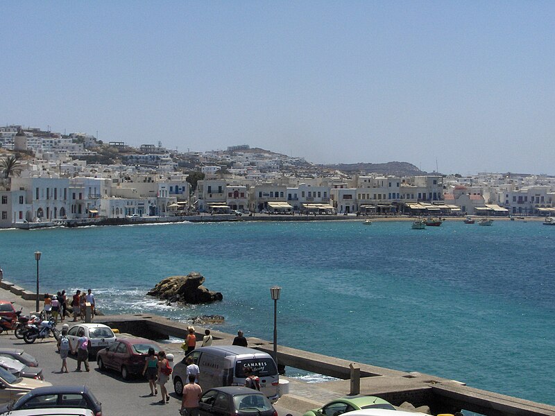 A ilha de Mykonos com prédios brancos ao longo de uma orla azul-turquesa. Pessoas e carros estacionados alinham a rua em primeiro plano. Representa aluguel de carros em Mykonos.