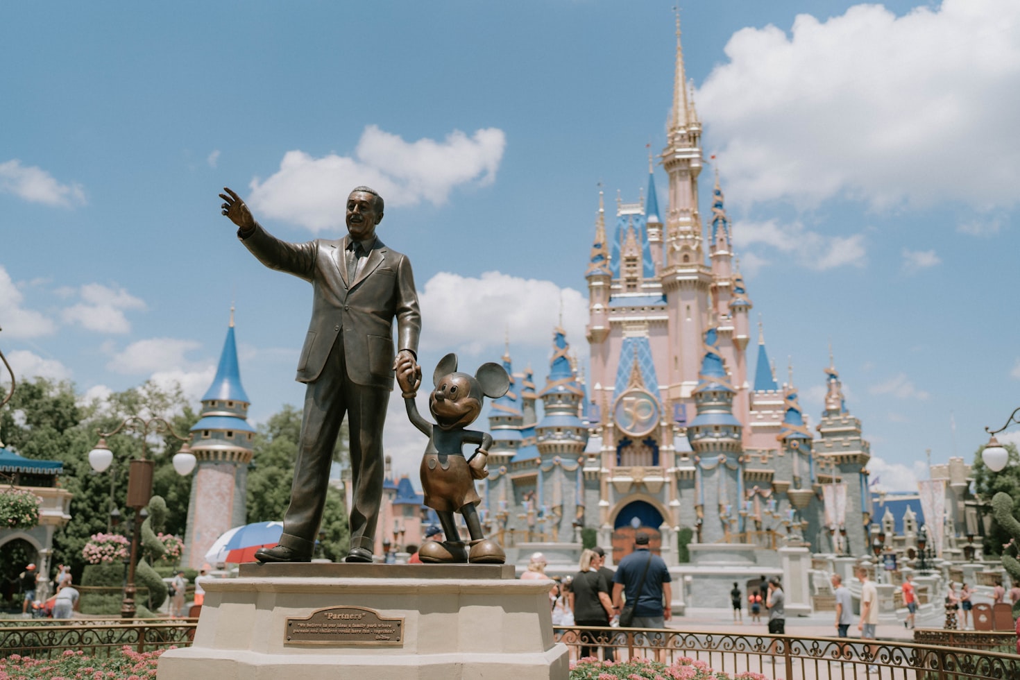 Estátua de um homem com um rato em frente a um castelo azul e dourado em um dia ensolarado, com pessoas ao redor, na Disney. Representa eSIM Disney.