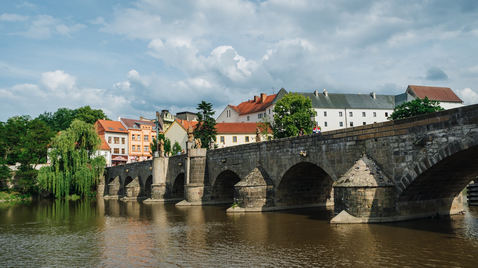 Ponte de pedra de de Písek com vários arcos sobre um rio, com edifícios coloridos e céu nublado ao fundo. Representa eSIM República Tcheca.
