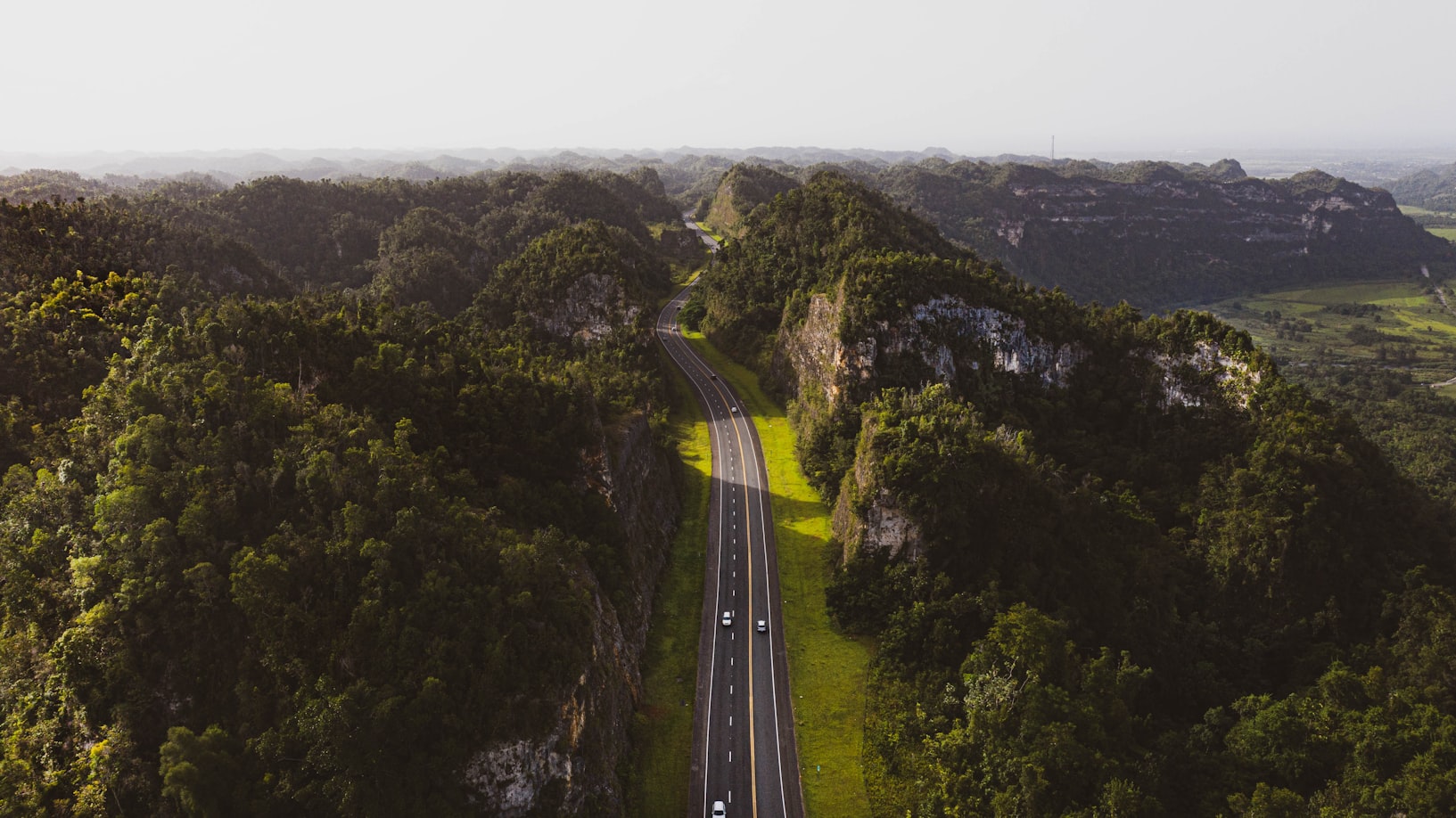 Vista aérea de uma rodovia em Porto Rico cortando um vale verdejante cercado por colinas, com vários veículos na estrada.