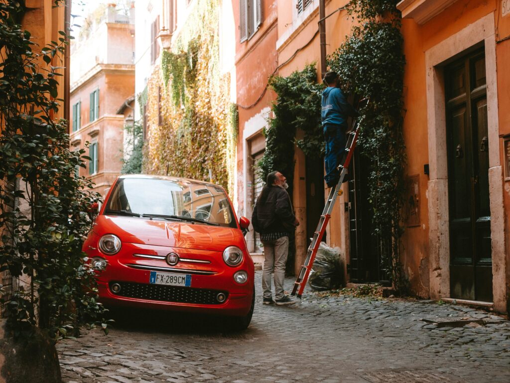 Um carro vermelho está estacionado em uma rua estreita de paralelepípedos. Dois indivíduos trabalham em uma escada, aparando plantas em uma parede de prédio.