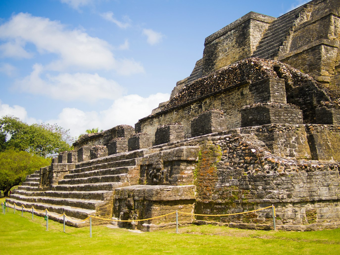 Uma estrutura piramidal de pedra com degraus e terraços sob um céu azul, cercada por grama e uma cerca de corda em primeiro plano, em Belize.