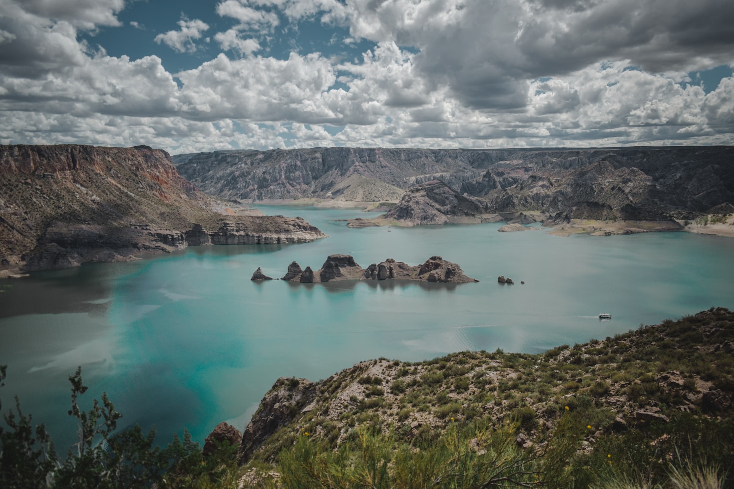 Um vasto lago azul-turquesa em Mendoza cercado por montanhas escarpadas sob um céu parcialmente nublado, com um barco visível na água. Representa esIM Mendoza.