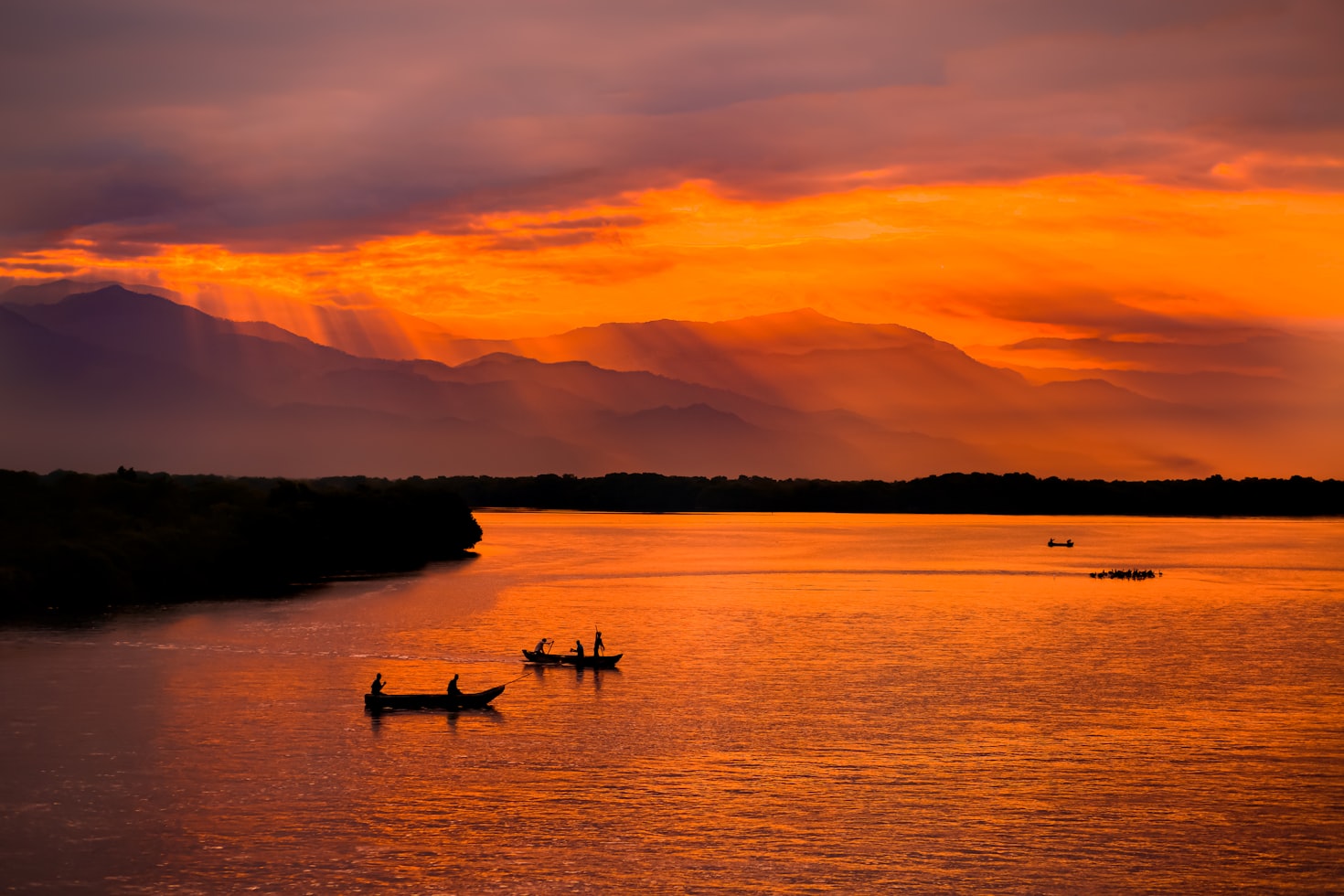 Barcos silhuetados em um rio calmo em Santa Marta com um pôr do sol alaranjado e raios de sol brilhando através das nuvens sobre montanhas distantes. Representa aluguel de carros na Colômbia.