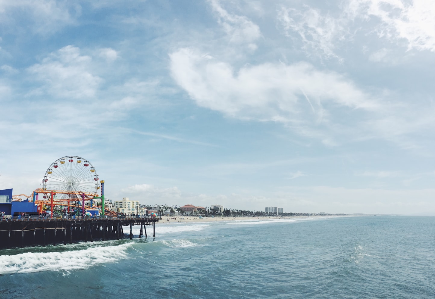 Uma roda gigante e um parque de diversões ficam no Pier de Santa Monica que se estende até o oceano, com uma praia de areia e o horizonte da cidade ao fundo. Representa eSIM Califórnia.