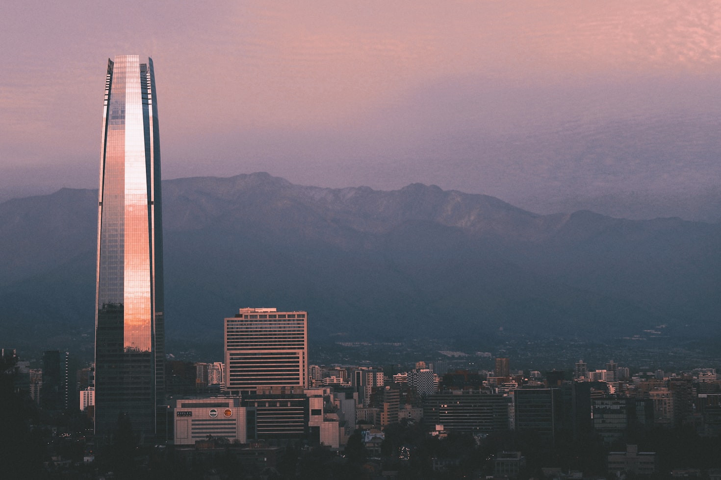 Arranha-céu alto refletindo a luz durante o pôr do sol, cercado por edifícios menores, com montanhas ao fundo sob um céu rosa, em Santiago. Representa eSIM Chile.
