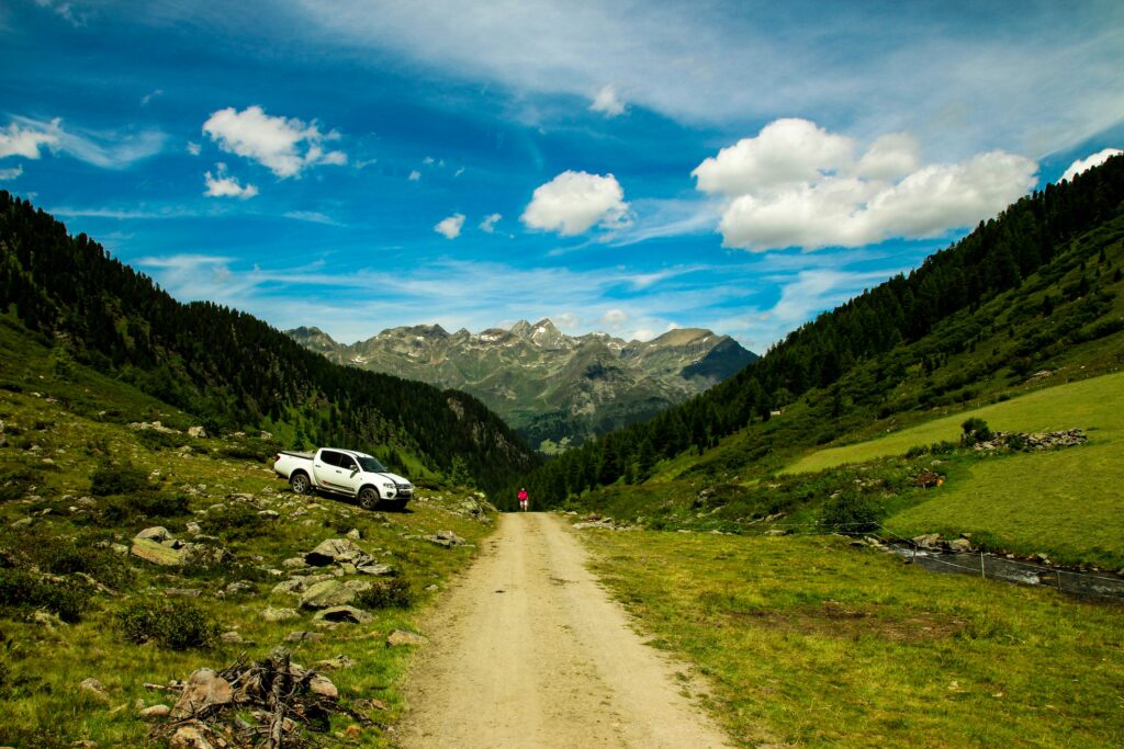 Um caminhão branco está estacionado ao lado de uma estrada de terra em um vale verdejante, com montanhas e um céu azul ao fundo, em See.