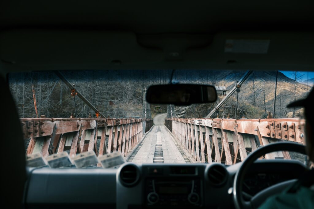 Vista de dentro de um veículo se aproximando de uma ponte pênsil estreita e desgastada, com tábuas de madeira, cercada por árvores nuas em Skippers Canyon Skippers Road. Representa aluguel de carros em Nova Zelândia.