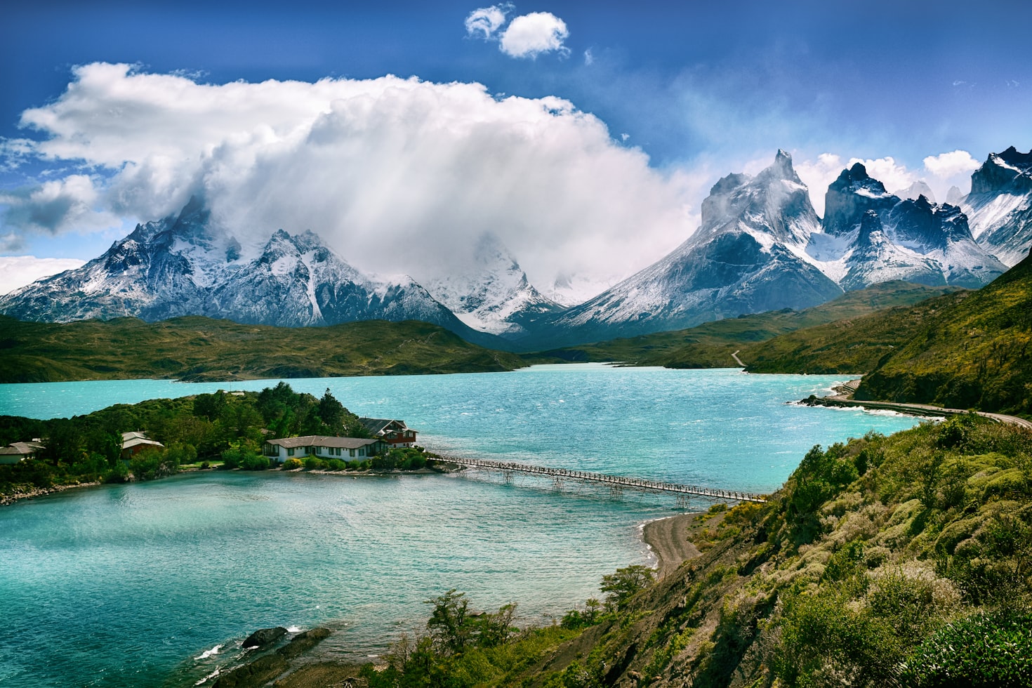 Lago turquesa com uma ponte de madeira, cercado por uma paisagem verde e montanhas cobertas de neve sob um céu azul parcialmente nublado, no Parque Nacional Torres del Paine. Representa eSIM Chile.