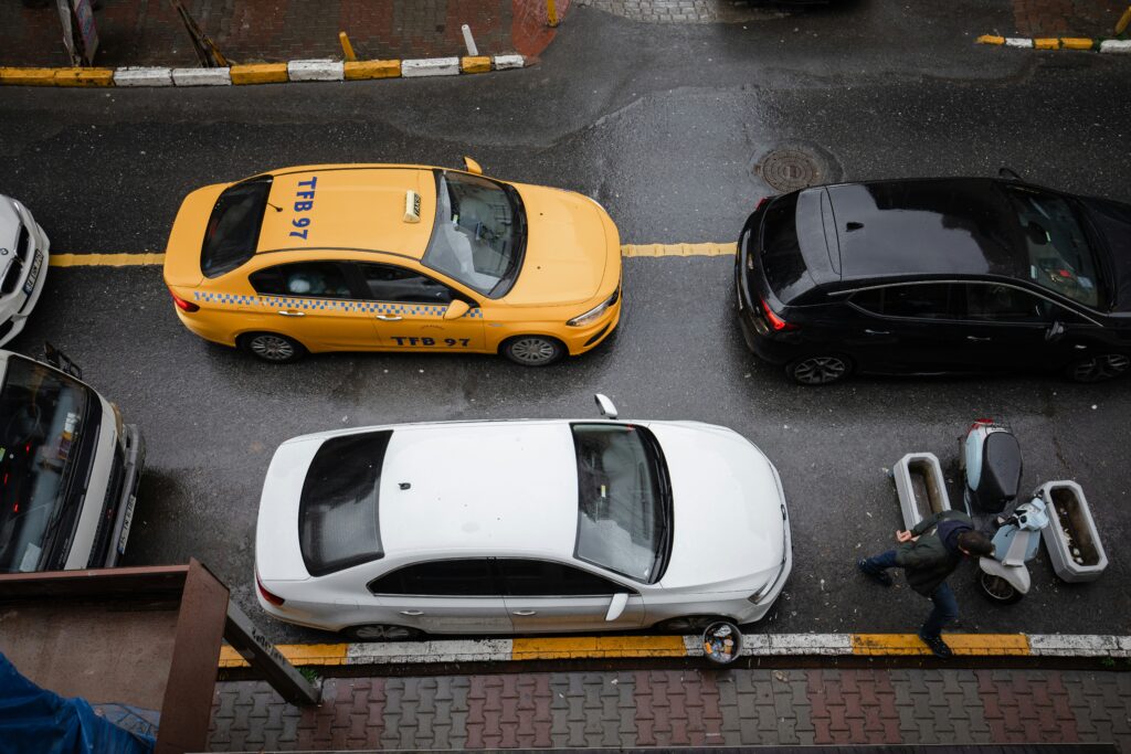 Vista de cima de um táxi amarelo, um carro preto e um carro branco parados em uma estrada de Istambul. Uma pessoa caminha perto na calçada molhada. Representa aluguel de carros em Istambul.
