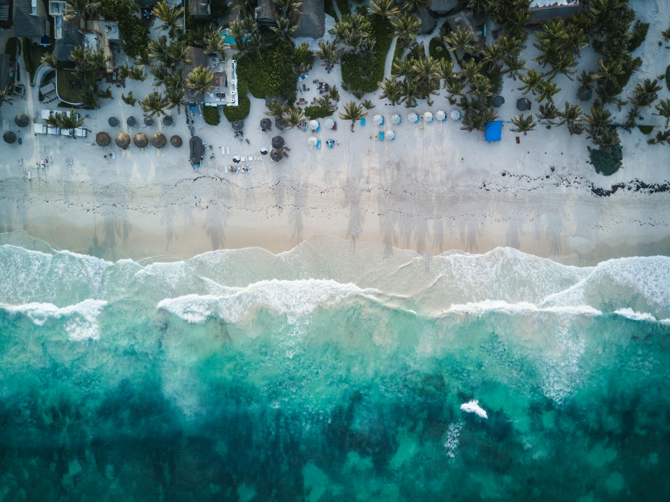 Vista aérea de uma praia em Tulum com águas azul-turquesa, palmeiras e guarda-sóis e espreguiçadeiras espalhados na areia branca. Representa eSIM México.