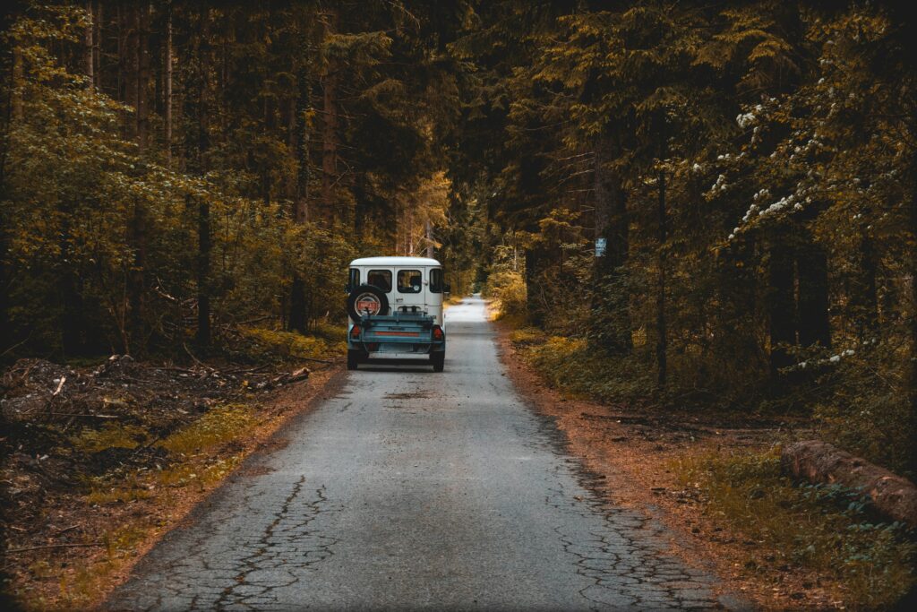 Uma van azul vintage passa por uma estrada estreita cercada por uma floresta densa, sob uma copa de árvores altas, em Vorarlberg. Representa aluguel de carros na Áustria.