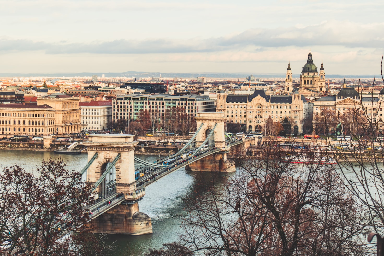 Vista de uma ponte suspensa sobre um rio em uma paisagem urbana com edifícios históricos e árvores em um dia claro,em Budapeste. Representa eSIM Budapeste.
