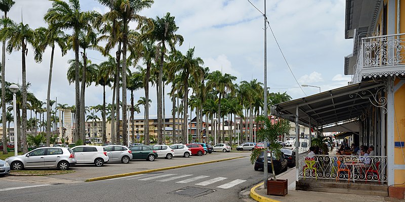 Cena de rua na Guiana Francesa com carros estacionados, palmeiras altas, faixa de pedestres, corrimão colorido e pessoas sentadas sob um toldo sombreado à direita. Representa Aluguel de carros na Guiana Francesa.