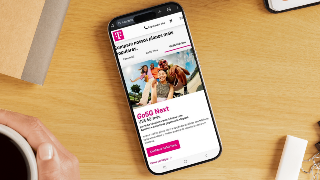 Um smartphone em uma mesa exibe o site do chip T-Mobile com um plano de celular. Uma mão segura uma xícara próxima.