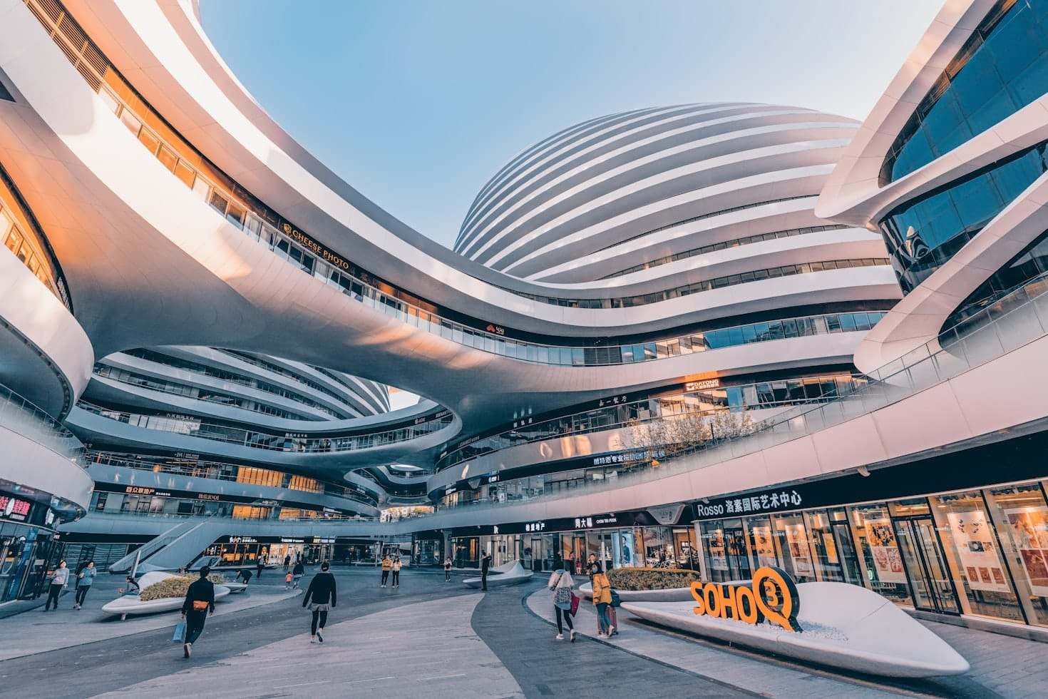 Edifício futurista de vários andares com superfícies brancas curvas e grandes passarelas abertas, com lojas e pessoas caminhando.