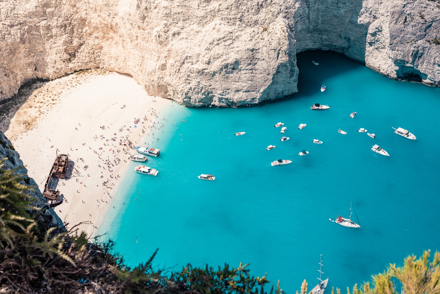 Vista aérea de uma praia com um naufrágio, areia branca e água azul cristalina; vários barcos estão ancorados nas proximidades, em Griekenland. Representa eSIM Zakynthos.