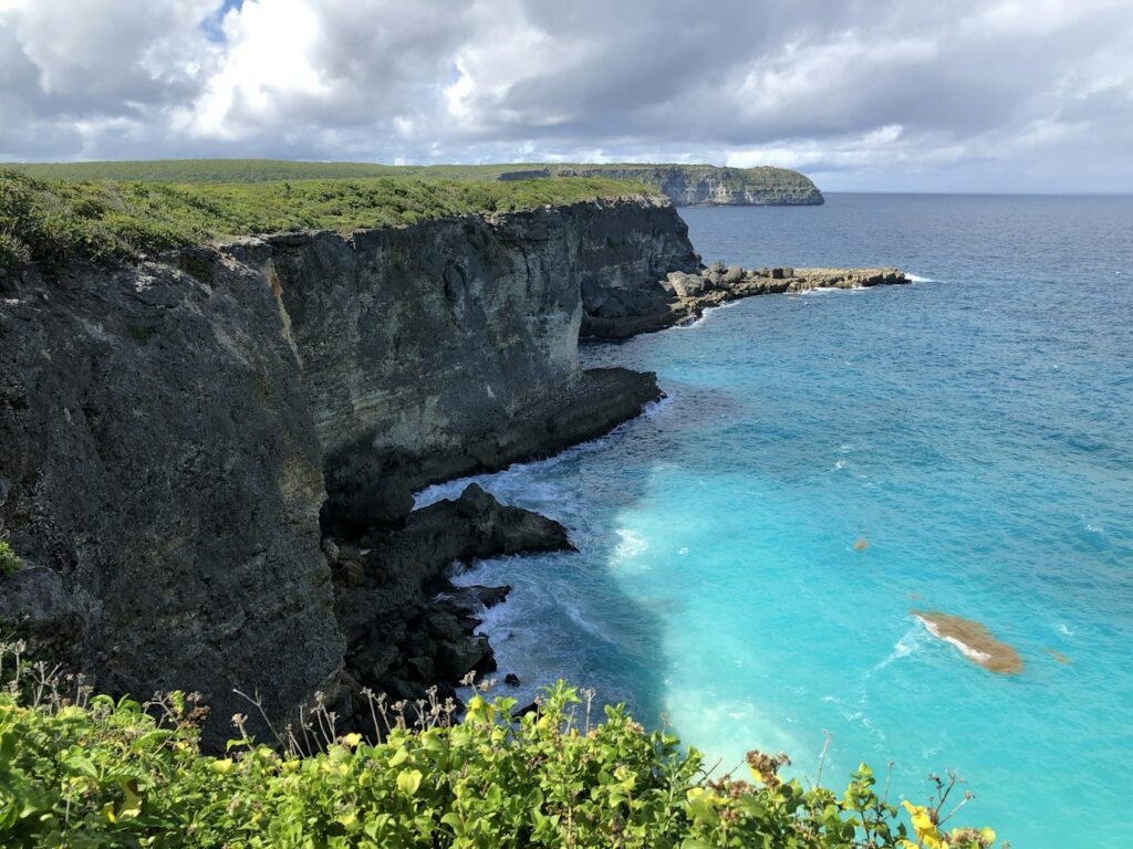 Penhascos rochosos têm vista para um oceano azul-turquesa sob um céu nublado, com vegetação exuberante no topo dos penhascos e ondas batendo na costa, em Grande-Terre. Representa eSIM Guadalupe.