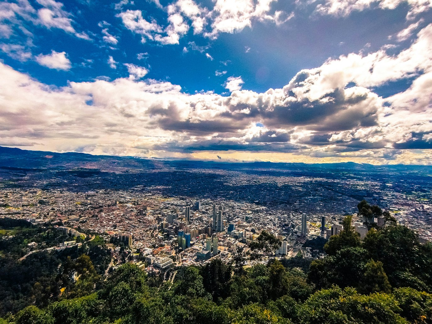 Vista panorâmica de uma paisagem urbana de Bogotá com edifícios altos, cercada por montanhas e sob um céu parcialmente nublado.