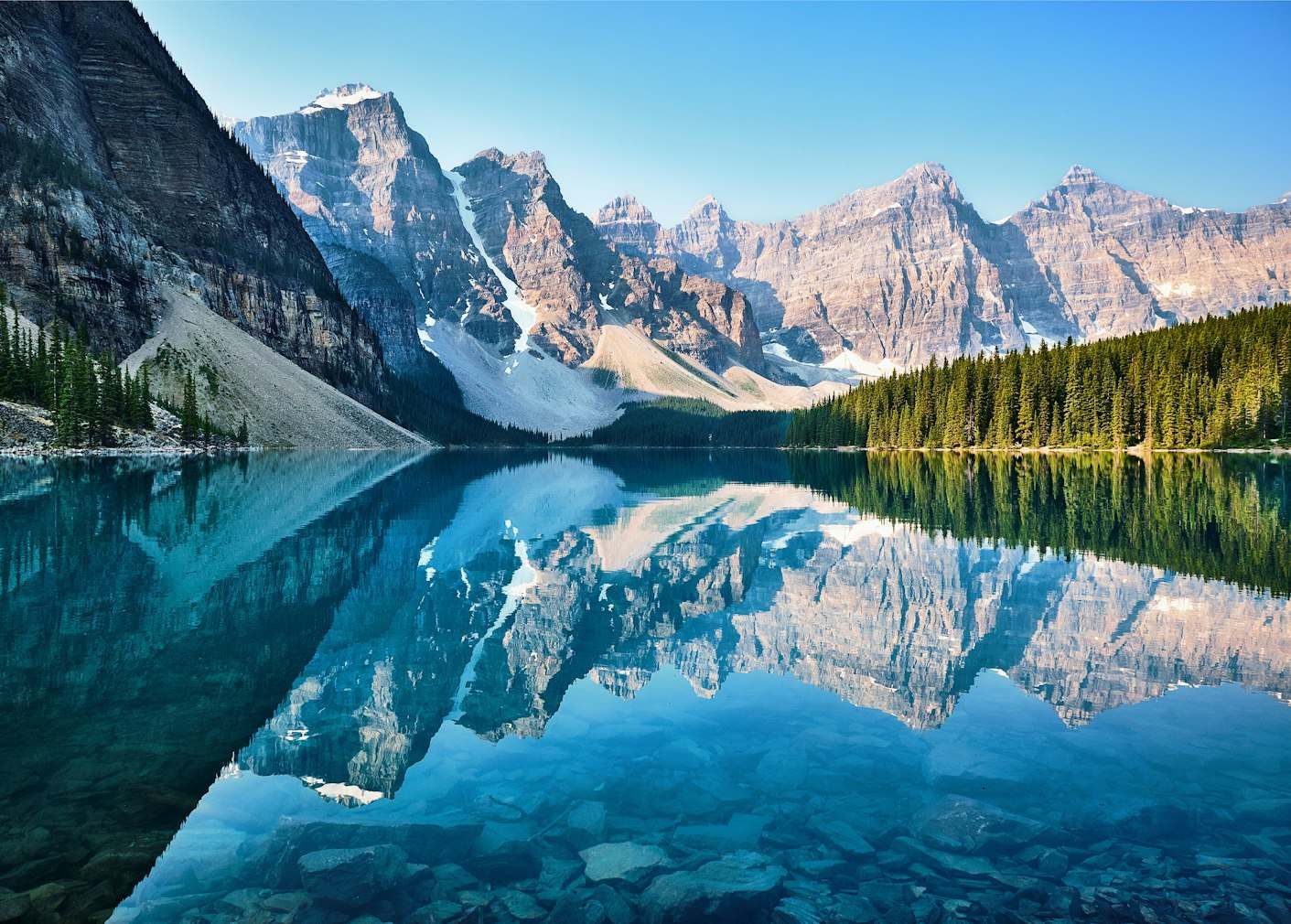 Um lago sereno reflete montanhas cobertas de neve sob um céu claro, com uma densa floresta perene ao longo da costa.