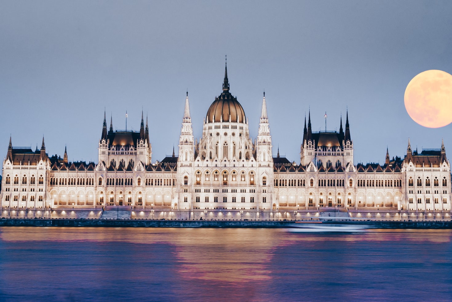 O edifício do Parlamento Húngaro iluminado à noite, refletido no Rio Danúbio, com uma grande lua cheia ao fundo. Representa eSIM Budapeste.