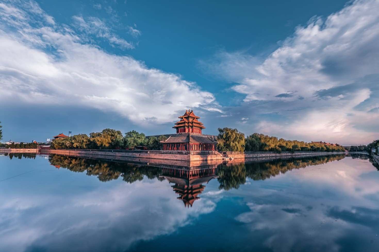 Pavilhão chinês tradicional cercado por árvores, refletido em águas calmas sob um céu azul com nuvens dispersas, em Pequim.