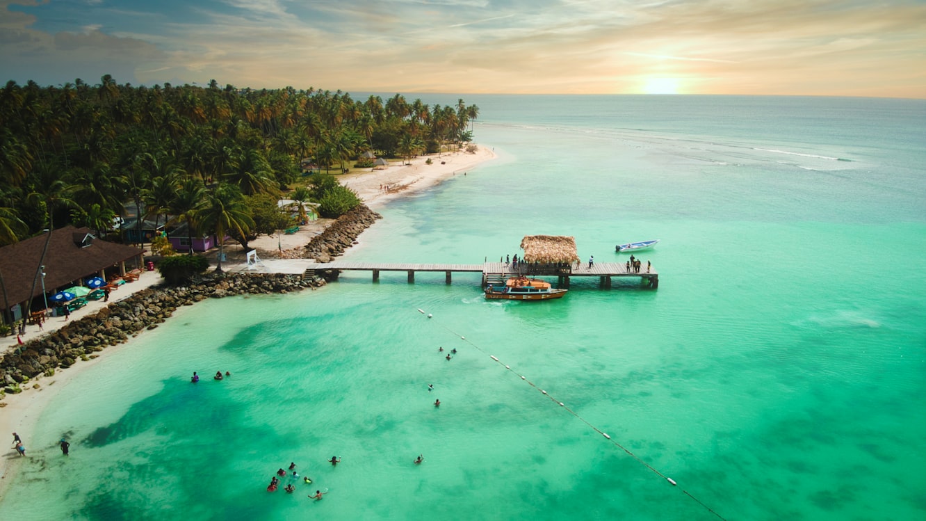 Vista aérea de uma praia tropical com palmeiras, um píer que se estende até águas azul-turquesa e pessoas nadando perto da costa. em Pigeon Point Beach. Representa Aluguel de carros em Trindade e Tobago.