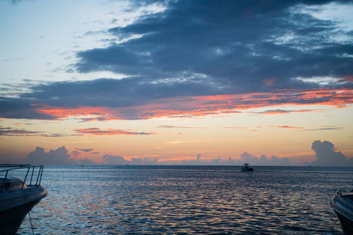 Pôr do sol nas Ilhas Cayman sobre um oceano calmo com nuvens dispersas. Um barco é visível à distância. Vistas parciais de barcos de cada lado. Representa aluguel de carros nas Ilhas Cayman.