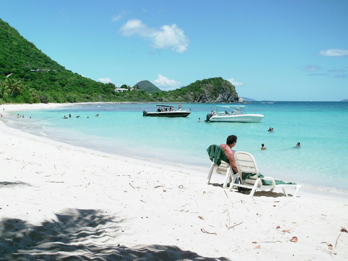 Pessoa sentada em uma espreguiçadeira em uma praia arenosa. Pessoas nadando em água turquesa, com barcos e colinas verdes ao fundo, em Tortola. Representa aluguel de carros nas Ilhas Virgens.
