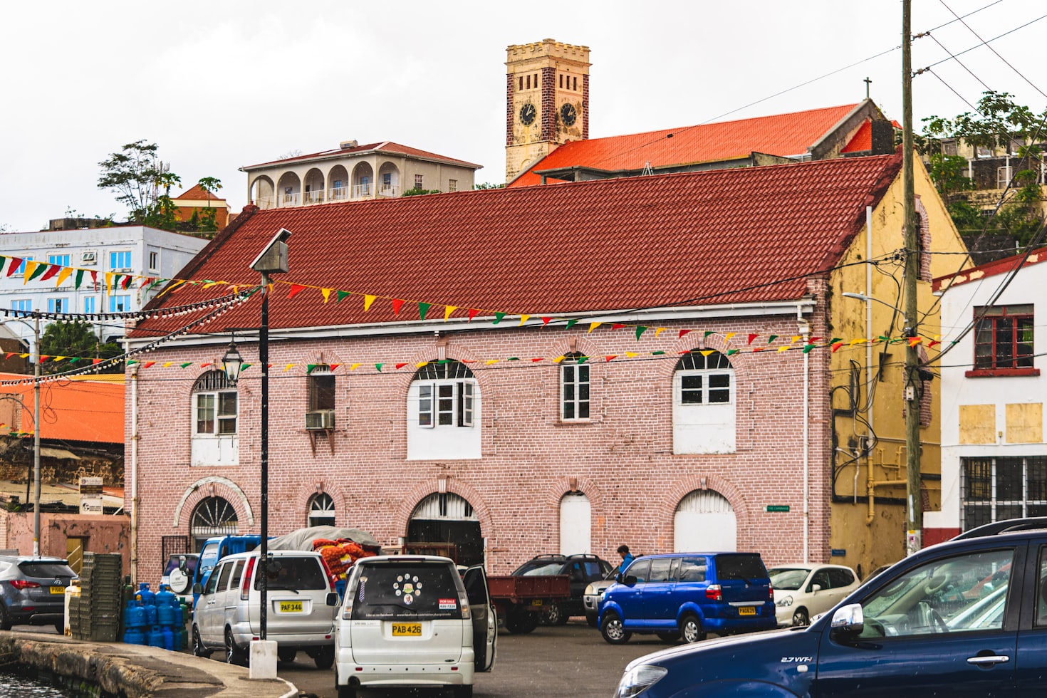 Vista da rua de um prédio de tijolos rosa com telhado vermelho, carros estacionados e bandeiras coloridas penduradas; torre do relógio ao fundo, em Granada. Representa aluguel de carros no Caribe.